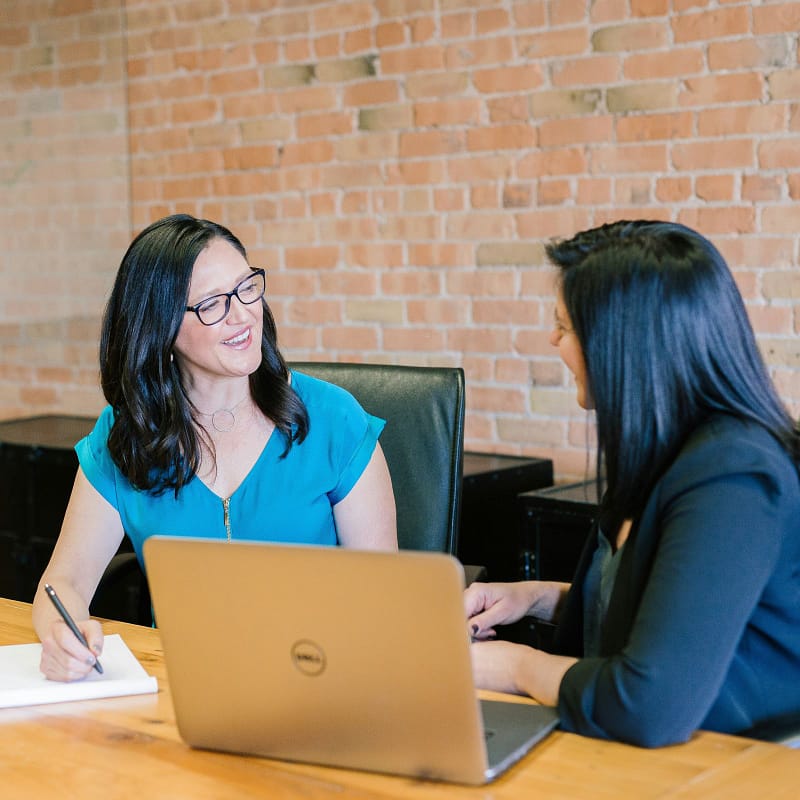 Two women having a friendly conversation at a desk in an office setting. One woman is taking notes on a notepad while the other is speaking. A laptop is open in front of them, and they are seated against a brick wall background.